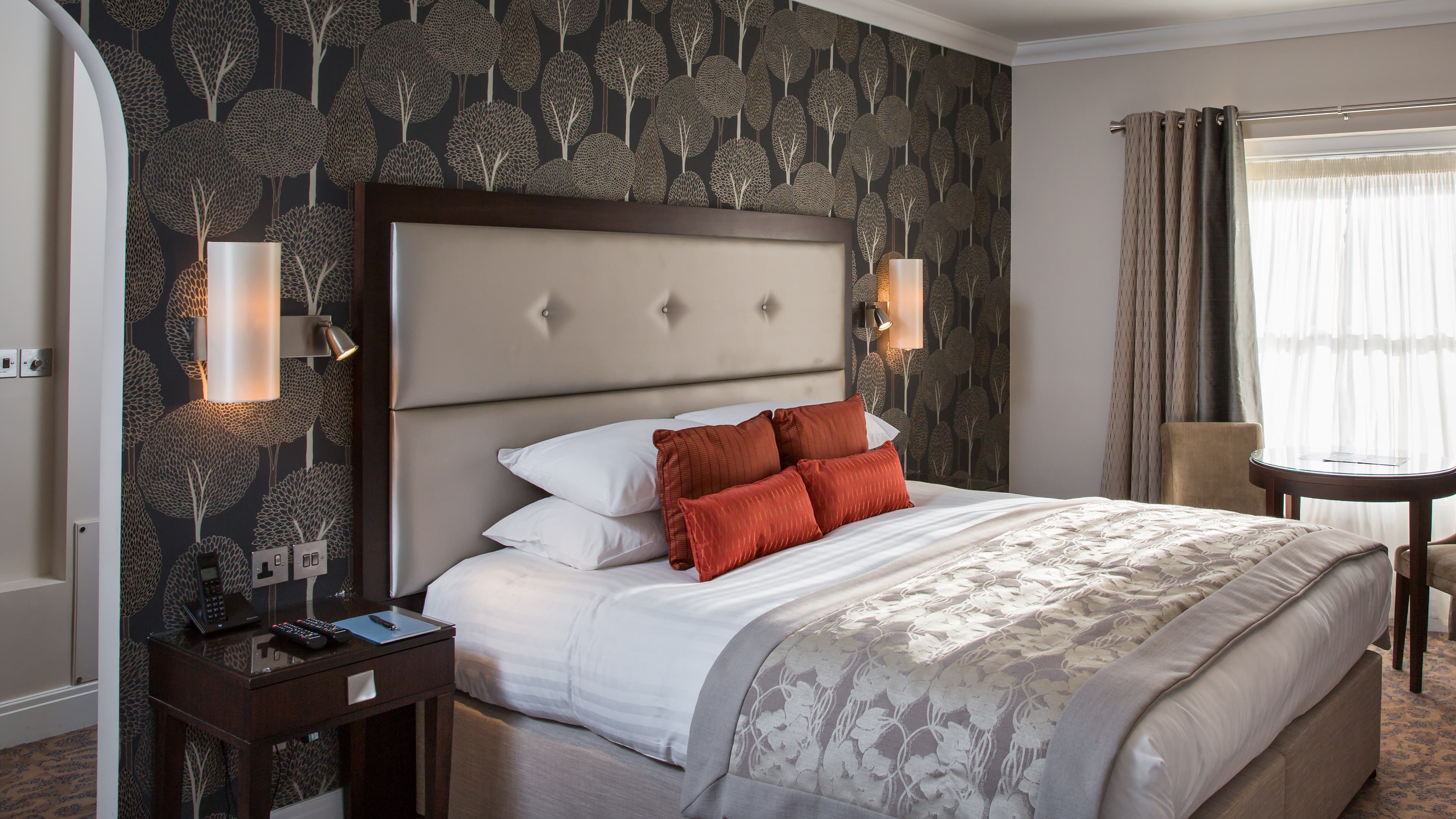 A photograph of a bedroom at Oakley Hall Hotel. The bed is luxurious with a silver throw and orange pillows on top of crisp white bedding.